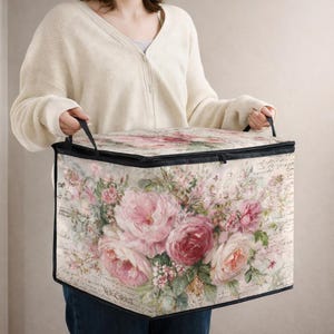 May include: A rectangular fabric storage bin featuring a vintage-inspired floral print with pink and red roses, green leaves, and cursive script text on a cream background. The container has black trim, a zippered lid, and two black fabric handles.