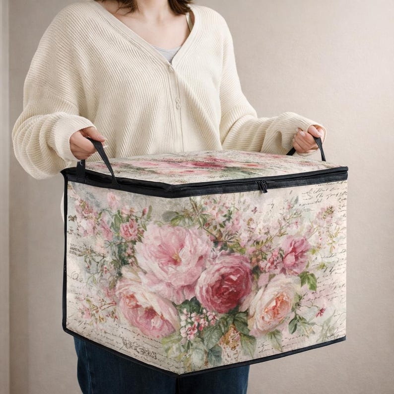 May include: A rectangular fabric storage bin featuring a vintage-inspired floral print with pink and red roses, green leaves, and cursive script text on a cream background. The container has black trim, a zippered lid, and two black fabric handles.