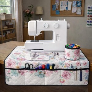 May include: A white sewing machine sits on a quilted mat featuring a floral pattern of pink roses and blue leaves. The mat includes multiple front pockets holding sewing tools and a matching cylindrical container filled with colorful thread spools.
