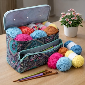 Op de afbeelding: Een paisley-patroon garen opbergtas met een lichtblauwe bovenkant, gevuld met kleurrijke garenballen. Verschillende garenballen en breinaalden zijn rond de tas gerangschikt. Een kleine potplant met roze bloemen staat in de buurt.