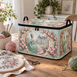 May include: A rectangular storage box with a light green background featuring a printed illustration of a person in a period gown standing before a large house, surrounded by pink roses and ornate gold frames, with black handles and a zipper closure.