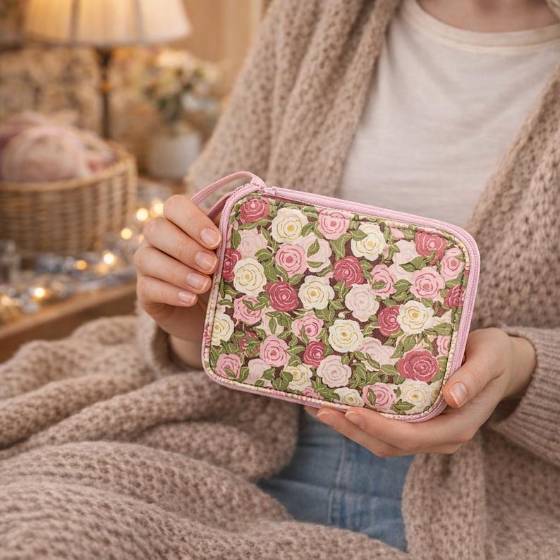 Pu&ograve; includere: Un piccolo astuccio rettangolare con stampa floreale, cerniera e manico rosa. L'astuccio presenta un motivo di rose rosa e bianche con foglie verdi su sfondo scuro. L'astuccio &egrave; tenuto in mano.