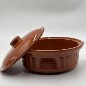 May include: A brown ceramic casserole dish with a lid. The dish has a round shape with a ribbed exterior and a lid with a small handle. The lid is slightly ajar, revealing the interior of the dish. The dish is designed for cooking and serving.