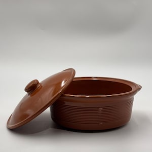 May include: A brown ceramic casserole dish with a lid. The dish has a round shape with a lid and a handle. The dish has horizontal lines around the exterior. The lid is slightly ajar.