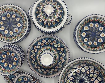 Może przedstawiać: Kolekcja białych ceramicznych naczyń do serwowania ze skomplikowanymi niebieskimi, zielonymi i czerwonymi wzorami geometrycznymi. Zestaw zawiera talerze, miski i naczynie do serwowania z pokrywką, wszystkie o pasującym wzorze. Krawędzie są wykończone kropkowaną obwódką.