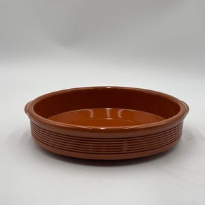 Puede incluir: Un plato redondo de cerámica de color terracota, de poca profundidad y con un borde ancho. El plato tiene una serie de estrías horizontales en el exterior y pequeñas asas. El interior es liso y brillante.