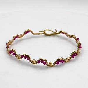 May include: A gold-coloured wire bracelet with a wavy design. The bracelet features small, round, fuchsia-coloured beads and clear, golden-coloured beads. It has a hook clasp. The jewellery is displayed on a white background.