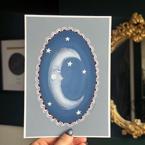 May include: A print depicting a crescent moon with a face, surrounded by stars, within a blue oval frame. The frame has a scalloped edge with a light brown and white border. The background is a light grey.