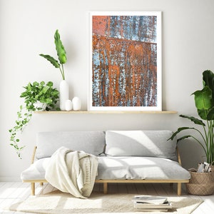 May include: A white couch with a throw blanket sits in front of a white wall with a framed abstract painting of rusty metal. The painting is on a wooden shelf with a potted plant on either side.