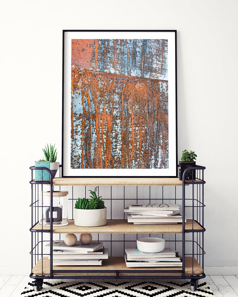 May include: A framed print of an abstract rusty metal surface with shades of orange, blue, and gray. The print is displayed on a black metal shelf with wooden shelves, books, and plants.