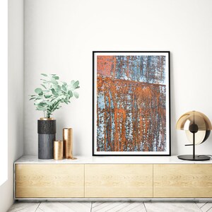 May include: A framed abstract art print featuring a rusty metal surface with blue and orange hues. The print is displayed on a light wood shelf with a vase and decorative objects.