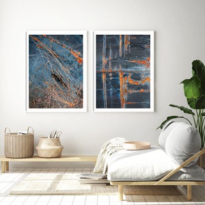 May include: Two framed abstract art prints with a blue and orange color scheme. The prints are hanging on a white wall above a wooden bench with a white cushion and a throw blanket.