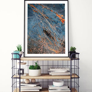 May include: A framed abstract art print with a blue background and orange and white lines. The print is displayed on a black metal shelf with wooden shelves. The shelf is filled with books, plants, and decorative items.