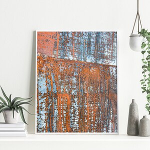 May include: Abstract art print featuring a close-up of rusty metal with blue and orange tones. The print is framed in a white frame and is hanging on a white wall.