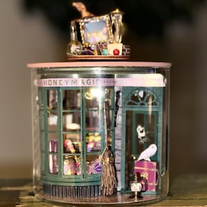 May include: A miniature Honeydukes shop diorama inside a glass jar. The shop has green trim, a detailed interior, and a small owl figurine. The jar sits atop a vintage book. A small decorative piece sits on top of the jar.