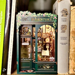 May include: A miniature green and brown "OWL BOOKSTORE" diorama with a detailed facade, including a "WELCOME" sign. The diorama is flanked by books, including a copy of "Oliver Twist". The bookstore features a detailed interior with miniature items.
