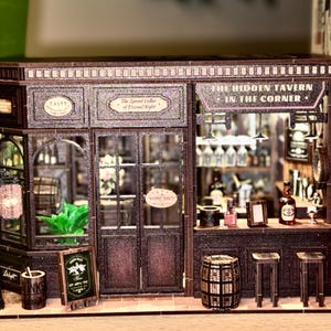 May include: A miniature tavern model with intricate details, including a sign that reads "The Hidden Tavern in the Corner." The model features a bar, stools, and a small chalkboard sign. The color scheme is primarily brown and dark wood.