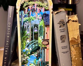 May include: A miniature book nook diorama featuring a garden scene with a blue arched bridge, a pond with lily pads, and pink wisteria hanging from above. A sign on the building reads Clos Normand. The structure is illuminated by small lights.