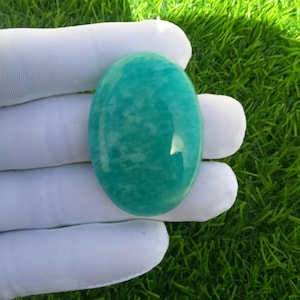 May include: A polished, oval-shaped gemstone in a vibrant teal color. The stone has a smooth, glossy surface with subtle internal patterns. The gemstone is held by a gloved hand against a backdrop of green grass.