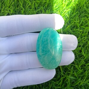 May include: A polished, oval-shaped Amazonite gemstone, showcasing a vibrant teal color with subtle white veining. The stone is held in a white gloved hand against a backdrop of green grass, highlighting its natural beauty.