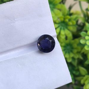 May include: A round, faceted gemstone with a deep, dark purple hue. The stone features a brilliant cut with multiple triangular facets that reflect light, creating a sparkling appearance. It is positioned on a plain, white, folded paper surface.