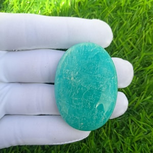May include: A polished, oval-shaped gemstone in a vibrant teal color. The stone has a smooth surface with subtle natural patterns and a slightly textured appearance. The gemstone is held in a white glove against a backdrop of green grass.