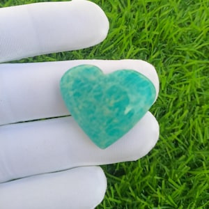 May include: A heart-shaped gemstone in shades of teal and turquoise. The polished stone has a textured appearance and is held by a white gloved hand. The background is green grass.