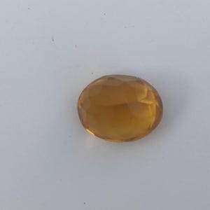 May include: A faceted, oval-shaped gemstone with a warm, amber hue. The gem is set against a plain white background, highlighting its clarity and color. The cut of the stone creates multiple reflective surfaces.