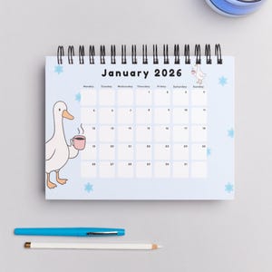 May include: A spiral-bound desk calendar for January 2026, featuring a light blue background with cartoon geese and star accents. The calendar includes a pen and pencil.