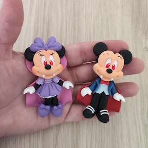Könnte beinhalten: Zwei gummiartige Figuren, die ein Vampir-Maus-Paar darstellen. Die linke Figur trägt ein violettes Kleid und einen Umhang mit passender Schleife. Die rechte Figur trägt ein blaues Smoking-Sakko mit roter Fliege und einem roten Umhang.