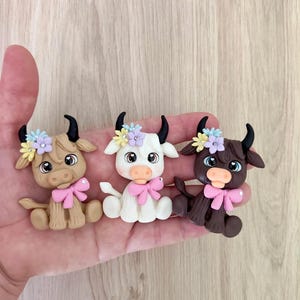 May include: Three small, sculpted animal figurines in light brown, white, and dark brown. Each figure features black horns, large expressive eyes, a pink bow at the neck, and a cluster of small yellow, blue, and purple flowers between the horns.