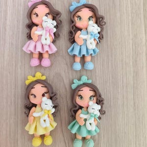 May include: Four decorative figurines of girls with brown curly hair, each wearing a solid-colored dress and matching hair bow in pink, blue, yellow, or mint green. Each figurine holds a small white teddy bear with a matching colored ribbon bow.