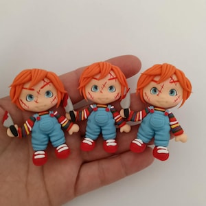 Könnte beinhalten: Drei handgefertigte Chucky-Puppen. Jede Puppe hat orangefarbenes Haar, blaue Augen und genähte Narben. Sie tragen blaue Latzhosen, rote Schuhe und gestreifte Hemden. Jede Puppe hält ein silbernes Messer.