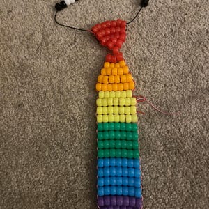 Könnte beinhalten: Eine handgefertigte Perlenkrawatte in Regenbogenfarben. Die Krawatte hat eine Halskette aus schwarzen und weißen Perlen, einen roten Perlenknoten und einen farbenfrohen Krawattenteil mit orangefarbenen, gelben, grünen, blauen und lila Perlen.