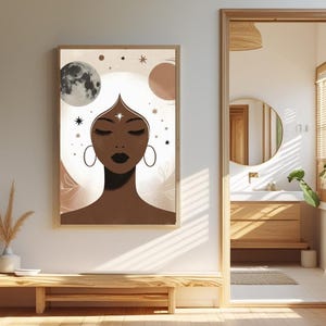 Op de afbeelding: Een ingelijste kunstdruk met een gestileerde illustratie van een vrouw met gesloten ogen, oorringen en een stervormig sieraad. Het kunstwerk bevat hemelse elementen zoals manen en sterren, in bruine, beige en crèmetinten. De print hangt aan een witte muur.