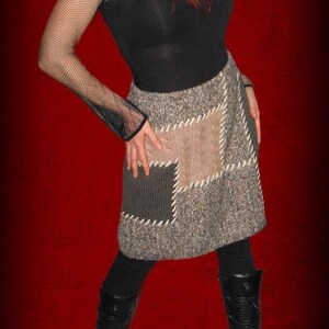 May include: A woman wearing a black top, a brown and grey patchwork skirt, black fishnet sleeves, a grey knit hat, and black boots. The woman has red and black hair.