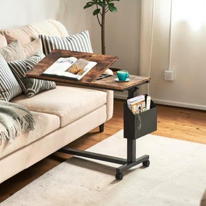 May include: A brown and black adjustable laptop table with a magazine holder. The table has a wooden top with a book and a teal cup on it. The table is next to a beige couch with pillows and a blanket.