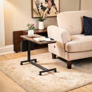 May include: A brown and black rolling laptop table with a wood-look surface. The table has a black metal frame and a small black storage compartment. A white teacup and saucer sit on the table, along with a magazine. The table is on a beige rug.