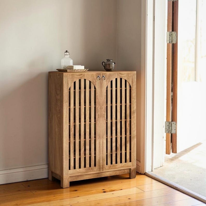 May include: A light brown wooden cabinet featuring two doors with vertical slats and arched tops. The cabinet has small circular metal pulls on each door. A clear glass bottle and a small dark ceramic vessel sit on the flat top surface.