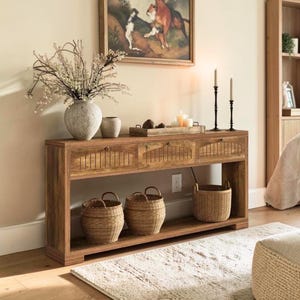 May include: A wooden console table with three drawers and a lower shelf, holding three woven baskets. The table is adorned with a vase of flowers, candles, and decorative items. A rug and artwork complete the scene.