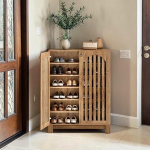 May include: A light brown wooden shoe cabinet with vertical slat doors, featuring six interior shelves filled with various pairs of shoes. The cabinet has one open door and one closed door, with a small woven basket and a plant placed on top.
