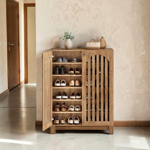 May include: A light brown wooden shoe cabinet with two doors featuring vertical slats and arched tops. The left door is open, revealing five interior shelves filled with various pairs of shoes. A small plant and two woven baskets sit on top.