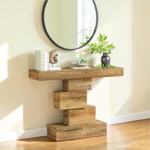 May include: A rectangular wooden console table featuring a unique stacked block base design. The table has a natural wood grain finish and a long, flat top surface. It is constructed from several horizontal wooden blocks arranged in a staggered pattern.