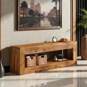 May include: A rectangular, light brown wooden console table with a framed design. The table has a lower shelf with two woven baskets, a small clock, a camera, and a bottle. A framed cityscape painting hangs above the table.