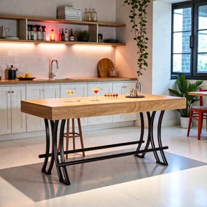 May include: A light-colored wooden kitchen island with black metal legs. The island has a rectangular top and is surrounded by a white kitchen with shelves, cabinets, and a window. Cocktails and bar tools are on the island.