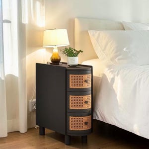 May include: A matte black bedside table with three curved drawers featuring natural light brown woven cane fronts and small dark knobs. A small lamp with a white shade and a potted green plant sit on the flat top surface of the narrow cabinet.