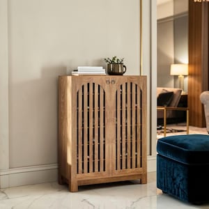 May include: A light brown wooden cabinet with two doors featuring vertical slats and circular metal pulls. The cabinet has a rectangular top surface holding a stack of white books and a dark green potted plant in a ceramic container.