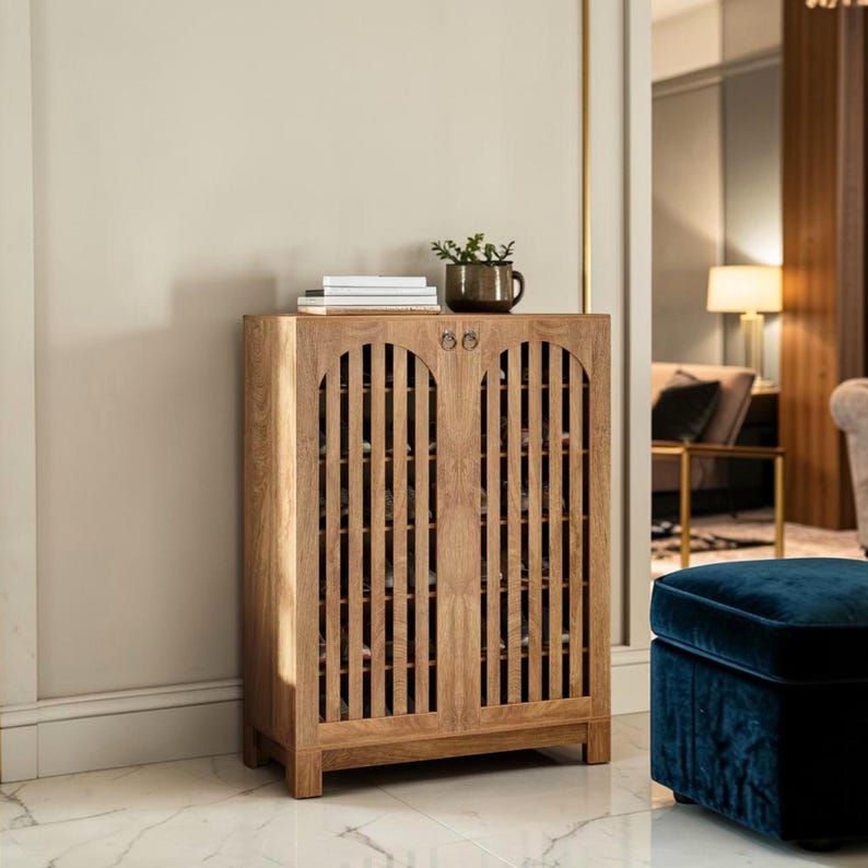 May include: A light brown wooden cabinet with two doors featuring vertical slats and circular metal pulls. The cabinet has a rectangular top surface holding a stack of white books and a dark green potted plant in a ceramic container.