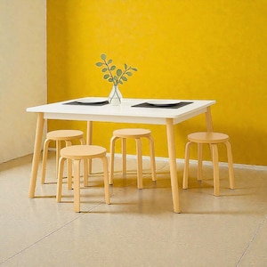 May include: A white rectangular dining table with four light-coloured wooden stools. The table has a white top and light-coloured legs. The stools are round with light-coloured wooden legs. A vase with green leaves sits on the table.