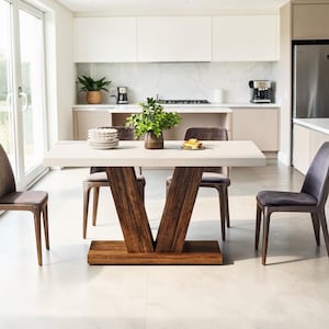 May include: A modern dining table with a light-colored rectangular top and a dark wood V-shaped base. Four gray upholstered chairs surround the table. A vase of greenery and a stack of plates are on the table. The room has a neutral color palette.